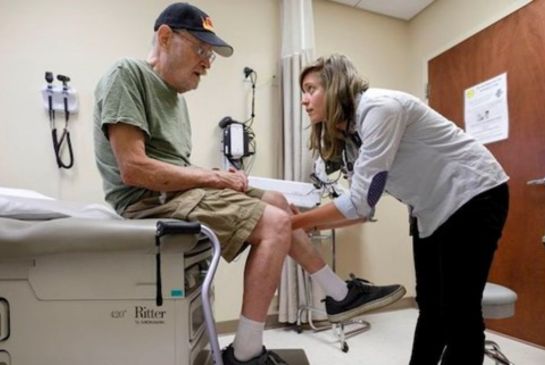 Doctor leans over and holds patient's knee as patient sits on exam table in an exam room.